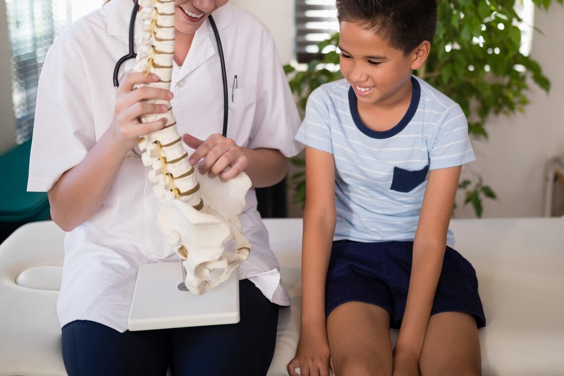 Smiling young female therapist explaining boy with artificial spine at hospital ward