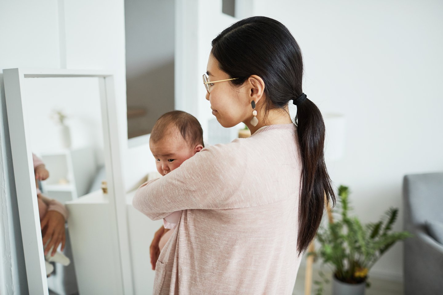 Rear view of young woman holding her baby and looking at mirror while standing in the room